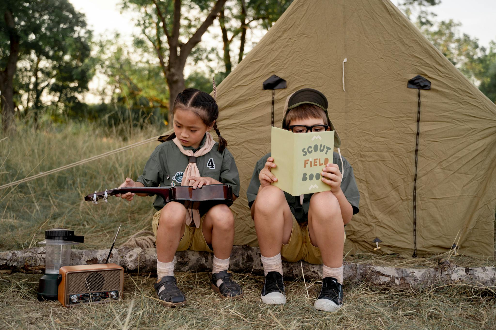 Children camping outdoors, playing ukulele and reading a scout book near a tent in nature.